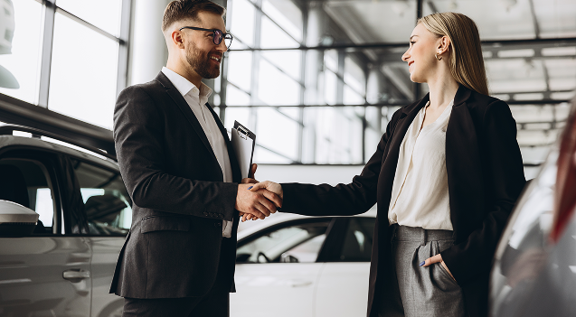 Man and woman shaking hands in a car dealership with cars in the background.
