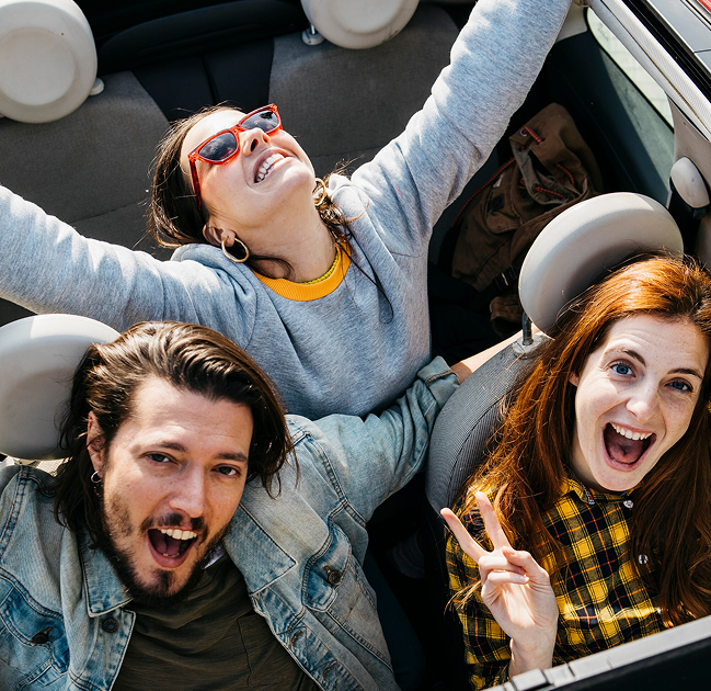 Three young adults smiling and looking up from inside a convertible car.